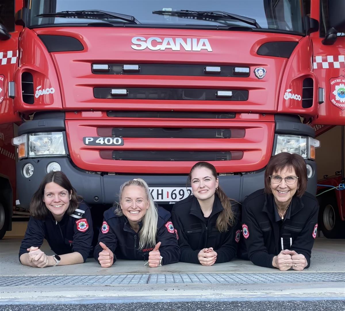Fire kvinnelige ansatte i brannvesenet som ligger under en brannbil og smiler til kamera - Klikk for stort bilde